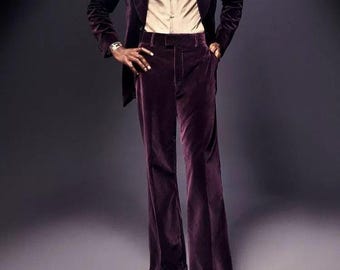 May include: A full-length shot of a person wearing a plum-colored velvet suit. The suit includes a blazer, flared trousers, and a button-down shirt. The shoes are a deep red color. The background is a dark gray.