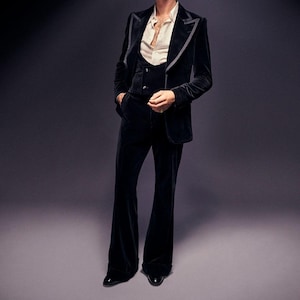 May include: A black velvet suit with a three-piece design. The suit includes a blazer with gray trim, a vest, and flared trousers. The outfit is paired with a cream-colored button-down shirt and black dress shoes. The background is a solid gray.