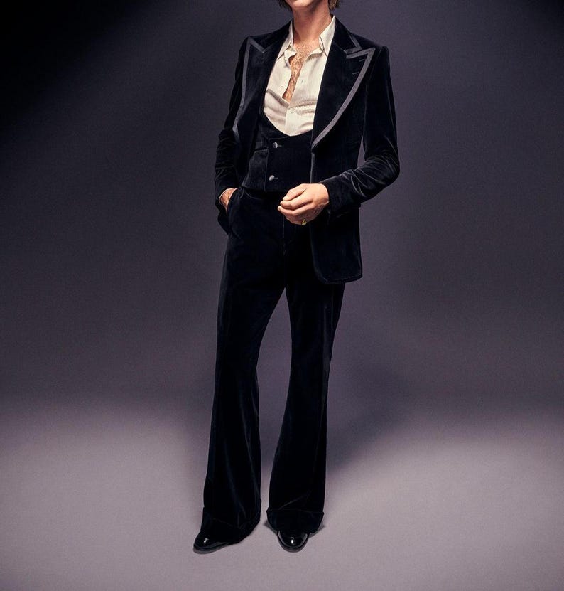 May include: A black velvet suit with a three-piece design. The suit includes a blazer with gray trim, a vest, and flared trousers. The outfit is paired with a cream-colored button-down shirt and black dress shoes. The background is a solid gray.
