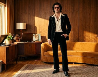 Puede incluir: Un hombre con un traje de terciopelo negro y gafas de sol está en una habitación de estilo retro. La habitación cuenta con un sofá color mostaza, paneles de madera y un tocadiscos vintage. La estética general recuerda a los años 70.