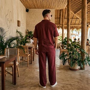K&ouml;nnte beinhalten: Ein bordeauxfarbenes Leinenhemd und ein passendes Hosen-Set. Das kurz&auml;rmelige Hemd hat einen Button-Down-Kragen. Die Hose hat ein gerades Bein. Das Outfit wird in einer tropischen Umgebung mit Holzm&ouml;beln und &uuml;ppigem Gr&uuml;n getragen.