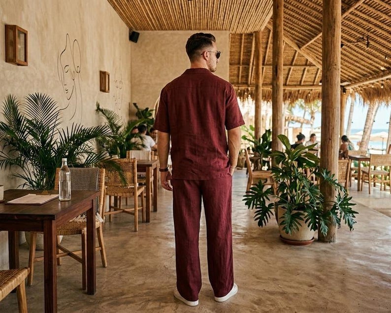 K&ouml;nnte beinhalten: Ein bordeauxfarbenes Leinenhemd und ein passendes Hosen-Set. Das kurz&auml;rmelige Hemd hat einen Button-Down-Kragen. Die Hose hat ein gerades Bein. Das Outfit wird in einer tropischen Umgebung mit Holzm&ouml;beln und &uuml;ppigem Gr&uuml;n getragen.