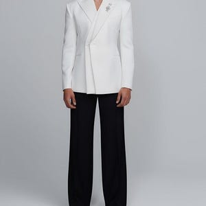 May include: A white blazer with a unique asymmetrical design, paired with black wide-leg trousers. The blazer features a decorative pin and a notched lapel. The outfit is a modern take on formal wear.