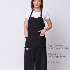 Può includere: Un grembiule nero indossato sopra una t-shirt bianca, dotato di due ampie tasche frontali e spalline regolabili. Il testo a destra recita: High degree of fabric breathability, Resistant to bleach splashes, Hair repellent. Il grembiule è lungo.