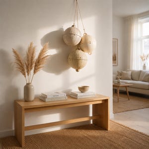 Può includere: Una panca in legno chiaro sostiene un vaso beige con erba della pampa essiccata, due libri impilati con l'etichetta DESIGN e una piccola ciotola. Tre ornamenti sferici in legno chiaro con motivi a griglia pendono dalla parete.