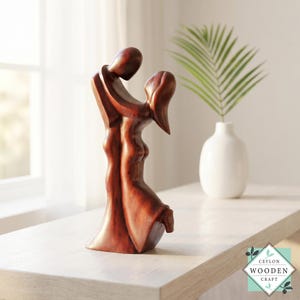 Puede incluir: Escultura de madera de un par de amantes abrazados, tallada en madera marrón rojiza. Las figuras están estilizadas con líneas suaves y fluidas, sugiriendo movimiento e intimidad. La escultura se exhibe sobre una superficie clara, con un jarrón blanco y vegetación al fondo.