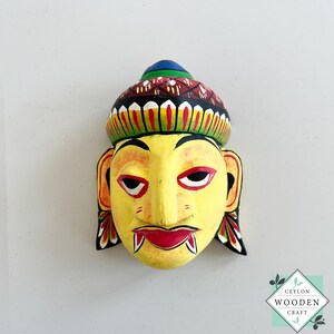 Könnte beinhalten: Eine handbemalte Holzmaske mit gelbem Gesicht, roten Lippen und schwarzen Augen. Die Maske hat einen dekorativen Kopfschmuck mit roten, grünen und blauen Akzenten. Die Maske ist ein dekorativer Gegenstand.