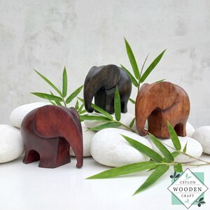 Puede incluir: Tres figuras de elefantes de madera en tonos marrones, desde caoba oscuro hasta roble claro. Los elefantes se exhiben sobre piedras blancas con hojas de bambú verdes, con el logotipo de Ceylon Wooden Craft.