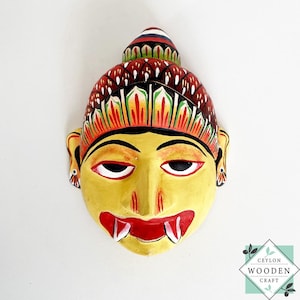 May include: A hand-painted wooden mask with a yellow face, red lips, and white fangs. The mask has a decorative headdress with red, green, and black accents. The mask is a traditional craft from Ceylon Wooden Craft.