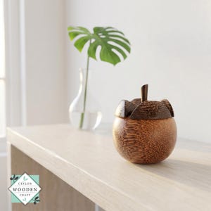 Op de afbeelding: Een ronde, gepolijste houten container met een donkerbruin, getextureerd deksel met een centraal steeltje. Het lichaam van de container toont een natuurlijk, lichtbruin houtnerfpatroon. Een logo met de tekst CEYLON WOODEN CRAFT is zichtbaar.