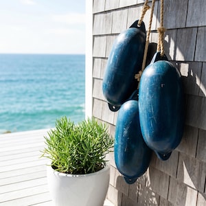 May include: Three weathered, dark blue cylindrical boat fenders hang from a thick, light-colored rope against a gray shingled wall. A vibrant green succulent plant sits in a smooth, white ceramic pot in the foreground, with the ocean visible.
