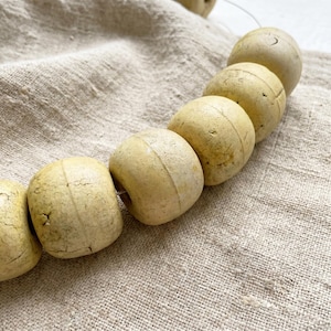 May include: A string of round, off-white beads with a matte, textured surface and visible cracks. Each bead features a central vertical line running around its circumference. The beads are arranged in a curved line, showing a weathered appearance.