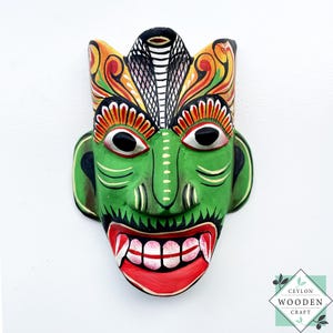 Puede incluir: Máscara de madera pintada a mano con una cara verde, boca roja y dientes blancos. La máscara presenta grandes ojos negros, un tocado tipo cobra y detalles decorativos en amarillo, naranja y rojo. Es un objeto decorativo.