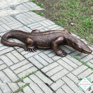 Puede incluir: Escultura detallada de color bronce de un lagarto, posiblemente un cocodrilo o un caimán, sobre una superficie de ladrillos. La escultura tiene escamas texturizadas y está colocada en diagonal.