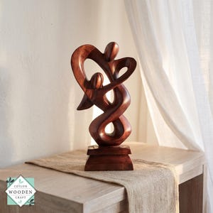 Puede incluir: Una escultura de madera de color marrón oscuro que representa dos figuras abstractas entrelazadas en una forma fluida y continua. La pieza presenta superficies lisas y pulidas y descansa sobre una base rectangular. El texto CEYLON WOODEN CRAFT es visible.