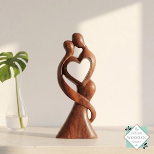 May include: A smooth, dark brown wooden sculpture depicting two abstract figures embracing to form a central heart-shaped negative space. The carving features a polished finish with visible wood grain patterns and stands on a wide, stable base.