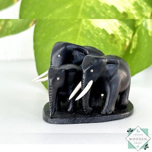 May include: A black wooden sculpture of three elephants with white tusks. The elephants are carved with detailed features and stand on a dark base. The sculpture is set against a background of green leaves and a white surface.