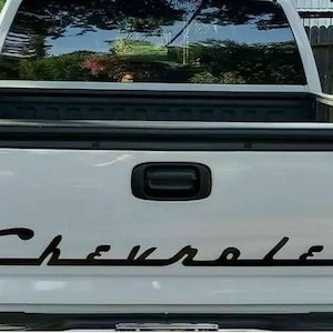 Puede incluir: Portón trasero de camioneta blanca con la palabra "Chevrolet" en negro en una fuente cursiva. La camioneta tiene luces traseras negras y un asa negra. La caja de la camioneta es visible.