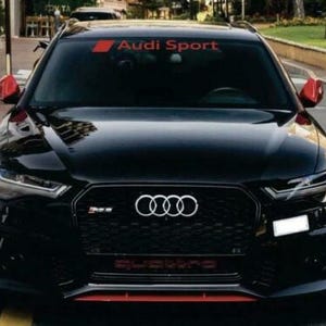 Op de afbeelding: Een zwarte Audi RS6 Avant met rode accenten, waaronder zijspiegels en een detail van de voorbumper. De auto heeft het Audi-logo en de tekst "Audi Sport" op de voorruit. Het woord "quattro" staat op de grille.