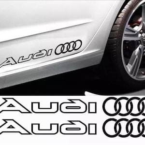 May include: Black Audi car decals. The decals feature the Audi logo and the word "Audi" in a stylized font. The decals are shown on a white car, near the tire.