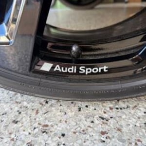 Puede incluir: Primer plano de una llanta de coche negra con el texto "Audi Sport" en blanco. La llanta tiene un acabado brillante y un tapón de válvula plateado. El neumático es negro y tiene un dibujo de la banda de rodadura visible. El fondo es una superficie gris moteada.