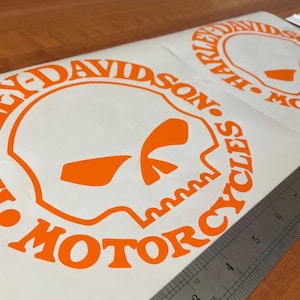 May include: Two orange Harley-Davidson Motorcycles decals on a white background. The decals feature a skull design within a circular frame, with the brand name and "MOTORCYCLES" text. A metal ruler is placed on the decals.