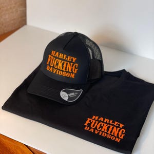 Pode incluir: Boné de caminhoneiro e camiseta pretos com texto laranja "HARLEY FUCKING DAVIDSON". O boné tem uma parte traseira em malha e uma viseira curva. A camiseta está dobrada, com o texto impresso na frente.