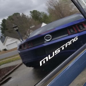 Op de afbeelding: Een blauwe Mustang-auto met het woord "MUSTANG" in witte letters op de zijkant. De auto staat op een natte straat, met huizen en bomen op de achtergrond.