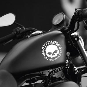 May include: Black and white image of a motorcycle gas tank with a Harley-Davidson logo. The logo features a skull within a circle, with the words "HARLEY DAVIDSON MOTORCYCLES" around it. The motorcycle's handlebars and mirror are visible.