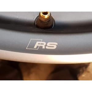 May include: Close-up of a section of a dark gray car wheel rim with the white letters "RS" inside a white square. A gold-colored valve stem is visible at the top.