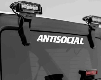 May include: Black vehicle rear window with the word "ANTISOCIAL" in white lettering. Two black LED light bars are mounted on the top of the window. The lights are attached with black brackets.