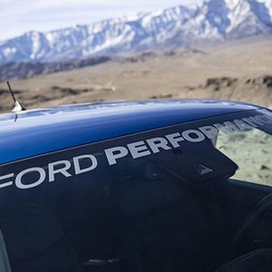 Könnte beinhalten: Ein blaues Auto mit dem Schriftzug "FORD PERFORMANCE" in weißen Buchstaben oben auf der Windschutzscheibe. Der Hintergrund zeigt eine Bergkette und eine Wüstenlandschaft.