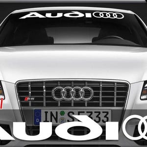 Op de afbeelding: Witte Audi auto met het Audi logo op de voorruit en de grille. Het woord "Audi" staat in het wit over de onderkant van de auto. De auto heeft heldere koplampen en een kentekenplaat.