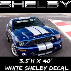 May include: A blue Shelby Mustang with white racing stripes. The windshield displays a white "Shelby" decal. The image also shows a white decal that is 3.5"H x 40" and says "WHITE SHELBY DECAL". The car is on a racetrack.
