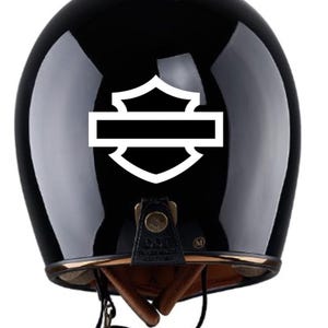 Könnte beinhalten: Glänzender schwarzer Motorradhelm mit weißem Emblem auf der Rückseite. Das Emblem hat die Form eines Schildes mit einem horizontalen Balken in der Mitte. Der Helm hat ein braunes Innenfutter und einen Kinnriemen.