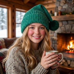 Può includere: Una donna con un berretto verde lavorato a maglia con un pompon, tiene in mano una tazza. Sorride, con un maglione accogliente. Un camino e una finestra innevata sullo sfondo creano una calda scena invernale.