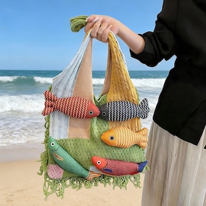 Può includere: Una colorata borsa tote con applicazioni a forma di pesce. La borsa presenta una base in rete verde, un manico intrecciato giallo e diversi pesci di peluche nelle tonalità del rosso, arancione e blu. Lo sfondo mostra una spiaggia e l'oceano.