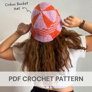 May include: A handmade crochet bucket hat in a vibrant color scheme of pink and orange. The hat features a striped pattern and is labeled "Cirkus Bucket Hat." The image also displays the text "PDF CROCHET PATTERN."