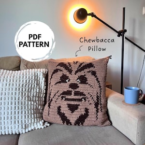 Può includere: Un cuscino all'uncinetto marrone con un disegno di Chewbacca. Il cuscino quadrato è appoggiato su un divano beige. Il testo "PDF PATTERN" è in un cerchio bianco. Le parole "Chewbacca Pillow" sono scritte sull'immagine.
