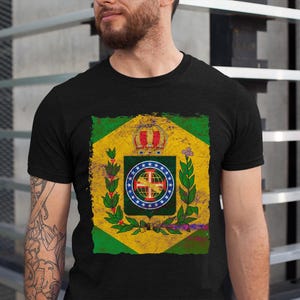 Könnte beinhalten: Schwarzes T-Shirt mit einem verblassten Grafik des brasilianischen Flaggenemblems. Das Emblem zeigt eine Krone, einen Globus und Lorbeerzweige auf gelbem und grünem Hintergrund. Das T-Shirt hat einen Rundhalsausschnitt.