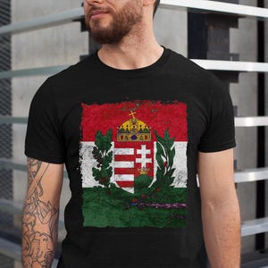 Könnte beinhalten: Schwarzes T-Shirt mit einem verblassten ungarischen Flaggen-Design. Die Flagge zeigt rote, weiße und grüne horizontale Streifen, eine Krone und ein Wappen. Das T-Shirt ist aus einem weichen Material gefertigt.