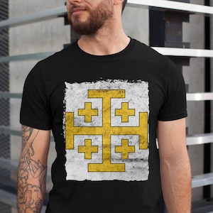 Könnte beinhalten: Schwarzes T-Shirt mit einem goldenen Jerusalemer Kreuz-Design auf weißem, verwittertem Hintergrund. Die Grafik ist zentriert. Das T-Shirt ist kurzärmlig und wahrscheinlich aus Baumwolle. Ein lässiges Kleidungsstück.