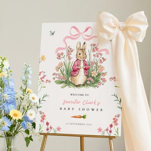 Puede incluir: Un letrero de bienvenida para baby shower con una ilustración de un conejito con una chaqueta rosa, rodeado de flores. El letrero dice "Bienvenida a la baby shower de Jennifer Clark" y la fecha, exhibido en un caballete dorado. Un gran lazo crema adorna el lado.