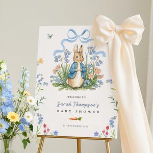 Op de afbeelding: Een babyborrelbord met een aquarel illustratie van Peter Konijn in een blauw jasje, omringd door bloemen. Het bord bevat de tekst "Welkom bij Sarah Thompson's Baby Shower" en de datum "15 september 2026". Het bord staat op een gouden ezel.