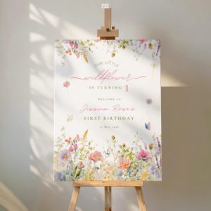 May include: A white sign on a wooden easel, featuring floral illustrations and the text "OUR LITTLE wildflower IS TURNING 1. WELCOME TO Jessica Rose's FIRST BIRTHDAY 21 May 2026". The design includes pink, purple, and yellow flowers.