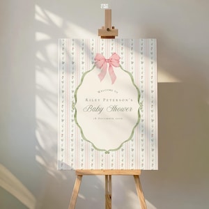 May include: A rectangular baby shower welcome sign featuring a pink and white vertical striped background with small floral accents. A central cream-colored scalloped frame displays the text Welcome to Riley Peterson's Baby Shower 18 December 2026.