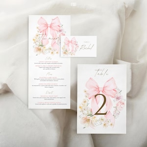 May include: Elegant wedding stationery set featuring a menu, place card, and table number. Each piece has a soft pink bow and floral design. The table number card displays the number 2 in gold.