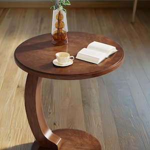 Pode incluir: Uma mesa lateral redonda de madeira marrom escuro com um suporte curvo. Um vaso com flores brancas, uma xícara e um pires e um livro aberto estão sobre a mesa. A mesa está em um piso de madeira.