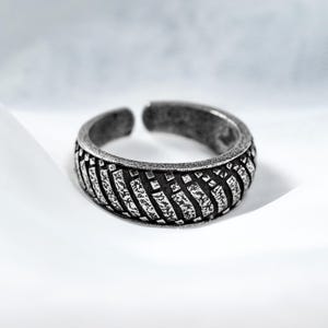 May include: A silver-toned adjustable ring with a textured tire tread design. The ring has an open design, allowing for size adjustment. The band features a repeating pattern of diagonal lines and rectangular shapes, creating a rugged, industrial aesthetic.