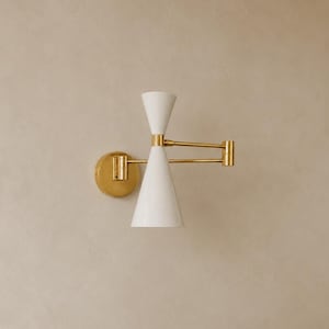 Op de afbeelding: Een wandlamp met een witte dubbelkegelvormige kap en een arm van gepolijst goudkleurig metaal. Het armatuur is voorzien van een ronde goudkleurige muurplaat en een verstelbaar zwenkarmmechanisme dat verbonden is met de zandlopervormige behuizing.